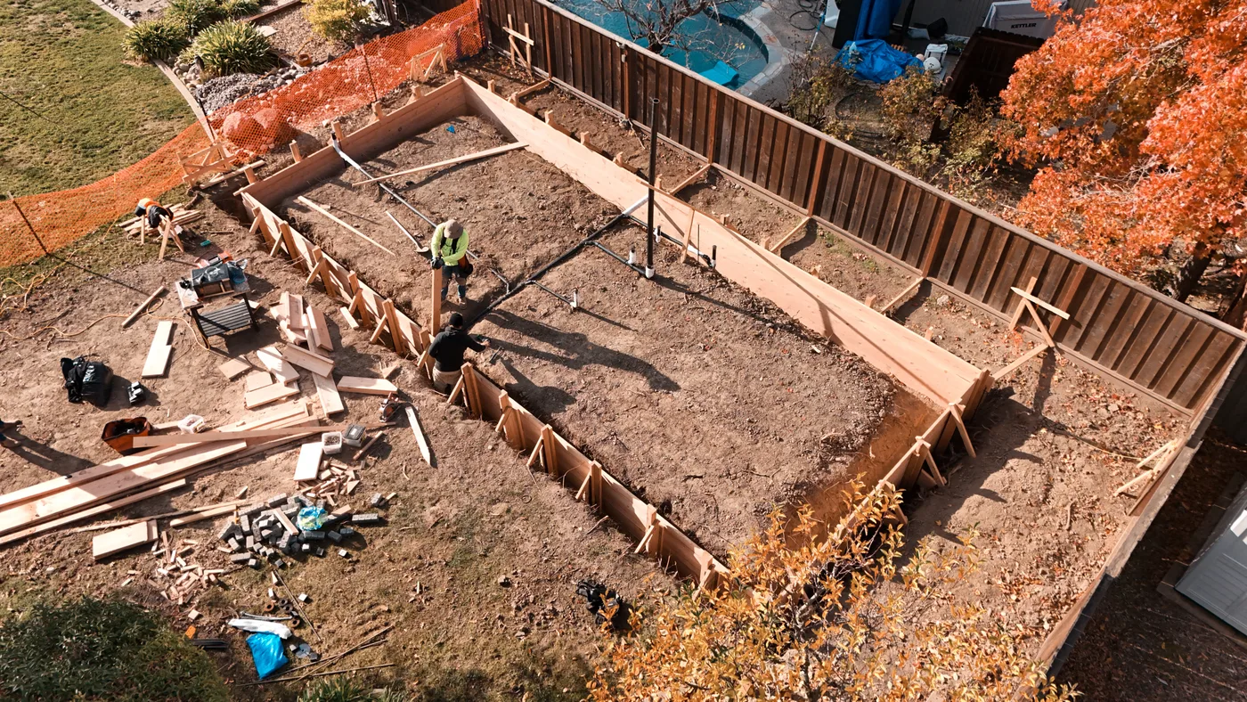 Crew setting the perimeter forms along the fence line, form lumber staged across the yard — Moraga Backyard ADU, Moraga, CA