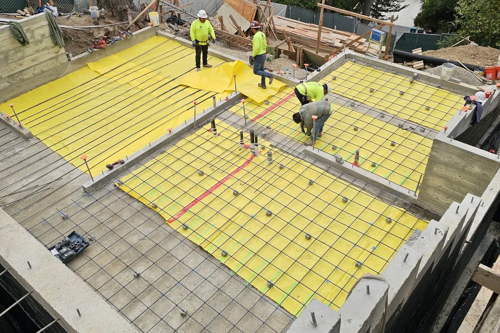 Foundation slab prep: yellow vapour barrier and a tied rebar mat inside poured concrete stem walls, with a crew working on site